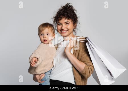 cheerful mother in casual clothes holding in arms toddler daughter and shopping bags isolated on grey,stock image Stock Photo