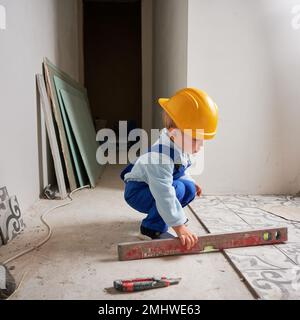 Child construction worker holding spirit level instrument. Kid in safety helmet and work overalls playing in apartment under renovation. Stock Photo