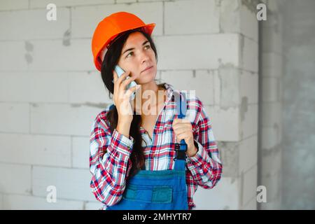 concentrated worker woman in blue overall having phone conversation during break from work at construction site indoors Stock Photo
