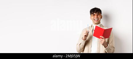 Cheerful young man in glasses laughing and taking notes, writing down in planner, holding pen and diary, standing on white background Stock Photo
