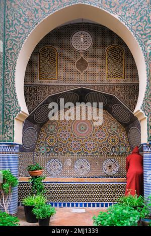 Fez, Morocco - man with red robe washes his hands in a water fountain in Fes el Bali. Monument with intricate mosaic tiles. Moroccan architecture. Geo Stock Photo