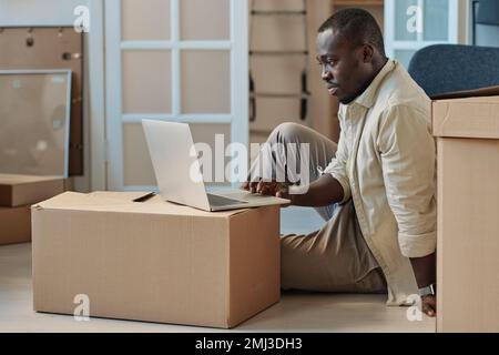 African American man sitting on the floor among packed boxes and searching for movers online using laptop Stock Photo