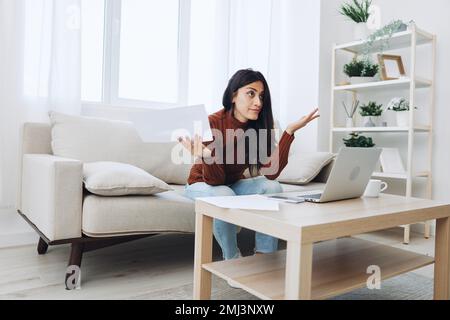 A sad freelance woman with a laptop on her desk works online from home shrugs her hands in incomprehension Stock Photo