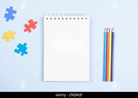 Blank open spiral paper notebook with blue, yellow, red pieces of puzzle and colored pencils on light blue background. World autism awareness day Stock Photo