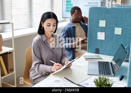 Portrait of young businesswoman writing on clipboard while working in office cubicle Stock Photo