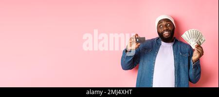 Dreamy african-american man showing credit card and dollars, thinking about shopping and smiling, standing over pink background Stock Photo