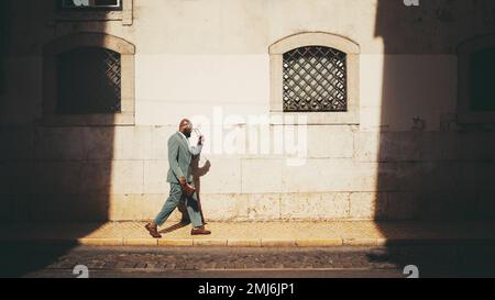 A side view of an dapper bald bearded black man entrepreneur in a green tailored summer suit passing by an illuminated part of the street next to the Stock Photo