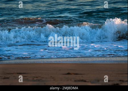The ocean's waves crash on a sandy beach at sunset. Stock Photo