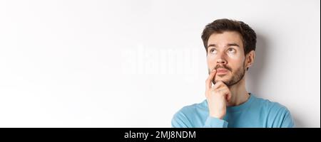Face of young caucasian man looking up pensive, making choice or thinking, pondering while standing over white background Stock Photo