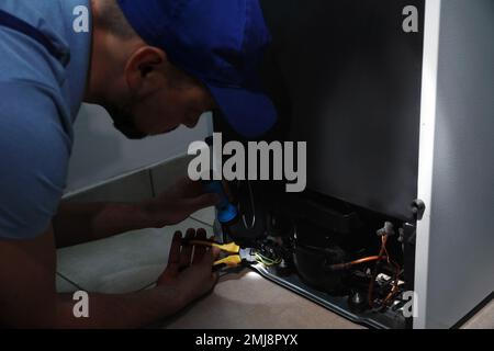 Professional electrician with flashlight fixing refrigerator indoors Stock Photo