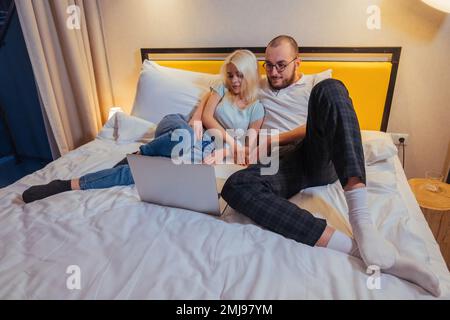 Happy relaxed young couple lying on sofa and watching film, photos or TV series on laptop. Stock Photo