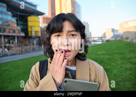 Close up portrait of girl with tablet, looks worried, concerned face expression, feels sad for someone, hears bad news, stands on street near Stock Photo
