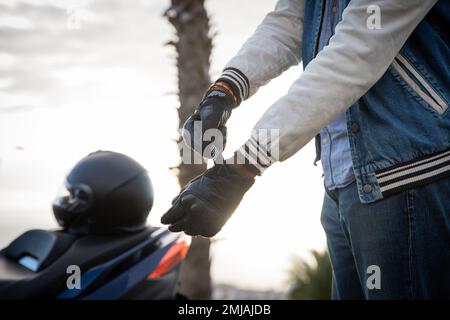 Hand of a biker putting on gloves with the scooter parked to the side with helmet on it Stock Photo