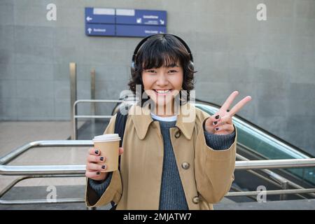 Positive korean girl in headphones, drinks coffee to go, shows peace, v-sign, stands on street and smiles happily Stock Photo