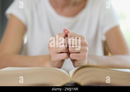 Woman sits at table with thick open book and holds her fists on book pages. Stock Photo