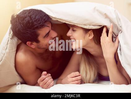 Keeping their love light and fun. a loving young couple smiling at each other while lying under a blanket together. Stock Photo