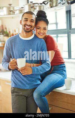 Starting our day with a lot of love. Cropped portrait of an attractive young woman embracing her husband while he drinks coffee in the kitchen. Stock Photo