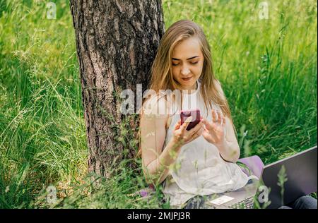 girl sits on the grass and works at a laptop. looks at the phone. freelance. selfeducation. the concept of remote learning and outdoor work Stock Photo