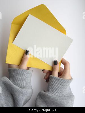 female hands in a sweatshirt with manicure holding a white sheet and a yellow envelope on a white background. postcard layout Stock Photo