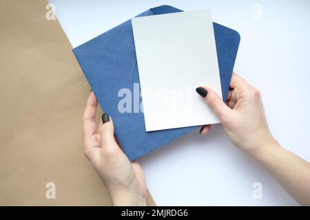 female hands with manicure holding a white sheet and a blue envelope vertically on a brown and white background.  postcard layout Stock Photo