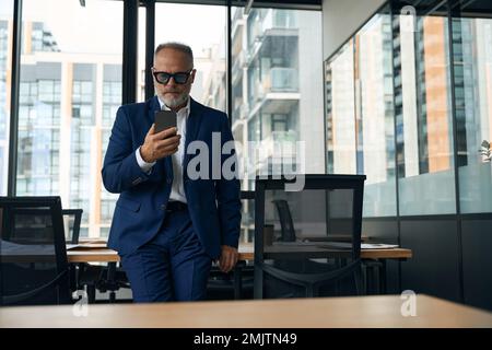 Focused supervisor in elegant suit looking at phone screen while standing in stylish modern office Stock Photo