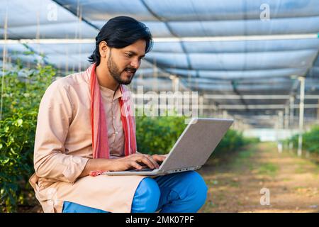 Happy young farmer busy working on laptop while sitting at greenhouse - concept of modern farming, technology and development or growth. Stock Photo