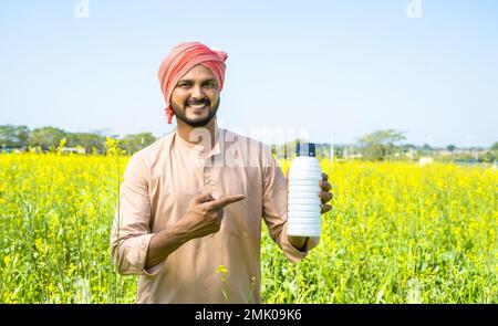 Confident young farmer showing fertilizer bottle by pointing finger while looking at camera at farmland - concept of product recommendation Stock Photo