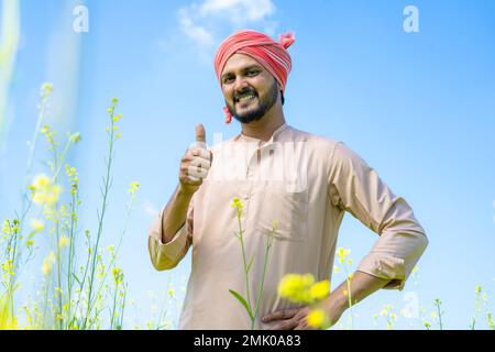 Young farmer standing with showing thumbs up by looking at camera at farmland - concept of successful, confident and modern agriculture Stock Photo
