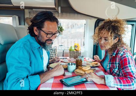 Couple man and woman inside a camper van planning next travel destination together using gps modern device on the table. Lunch time on vehicle motor h Stock Photo