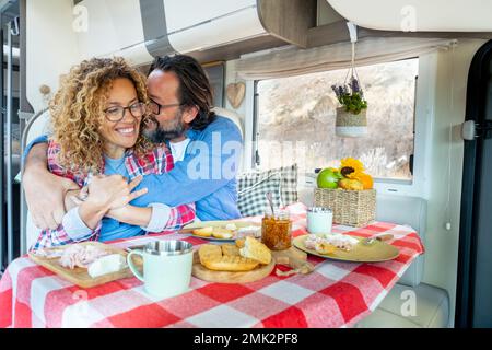 Excited and joyful couple in love inside camper van during lunch time. Cheerful woman and man hugging and having fun laughing during adventure travel Stock Photo