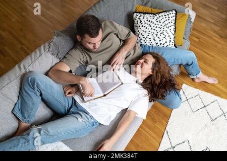 Couple in love lying down on sofa together reading book and smiling. Stock Photo