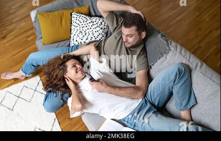 Couple lying on sofa in living room resting using mobile phone smiling, enjoying time together after work. Stock Photo