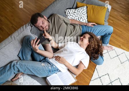 Playful couple lying down on sofa at home man using smart phone girlfriend touching boyfriends nose with her finger. Stock Photo
