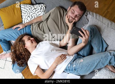 Happy couple resting on sofa at home girlfriend stretching boyfriend's mouth with her fingers making him smiling while man looking at screen of smart Stock Photo