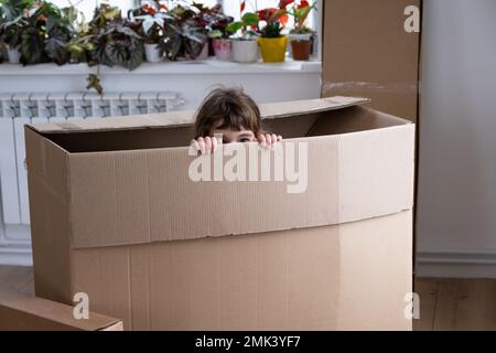 The girl hides in a big box and looks out of it funny in a room inside the home. Moving to a new house, unpacking things, renting an apartment, mortga Stock Photo