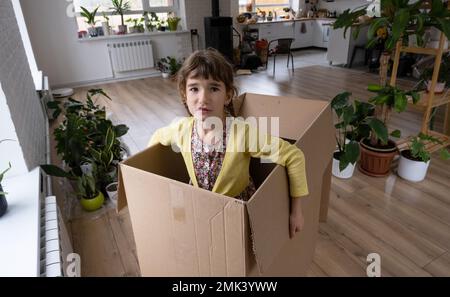 The girl hides in a big box and looks out of it funny in a room inside the home. Moving to a new house, unpacking things, renting an apartment, mortga Stock Photo