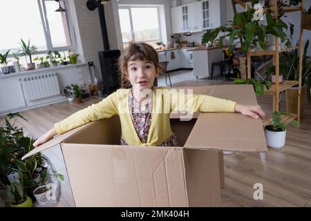 The girl hides in a big box and looks out of it funny in a room inside the home. Moving to a new house, unpacking things, renting an apartment, mortga Stock Photo