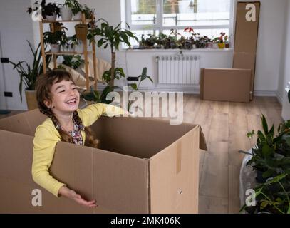 The girl hides in a big box and looks out of it funny in a room inside the home. Moving to a new house, unpacking things, renting an apartment, mortga Stock Photo