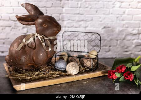 brown clay easter bunny in wooden bowl with easter eggs and hay in front of white brick wall with red tulips Stock Photo