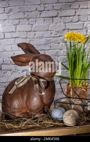 brown clay easter bunny in wooden bowl with easter eggs and hay in front of white brick wall with red tulips Stock Photo