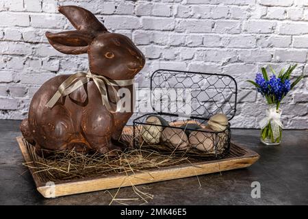brown clay easter bunny in wooden bowl with easter eggs and hay in front of white brick wall Stock Photo