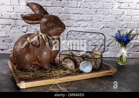 brown clay easter bunny in wooden bowl with easter eggs and hay in front of white brick wall Stock Photo