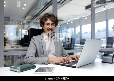 Portrait of hispanic businessman, man with curly hair and beard wearing glasses smiling and looking at camera, programmer typing on laptop inside bright office. Stock Photo