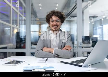 Portrait of successful financier businessman, hispanic man sitting at workplace working with documents and laptop inside office, man with crossed arms smiling and looking at camera. Stock Photo