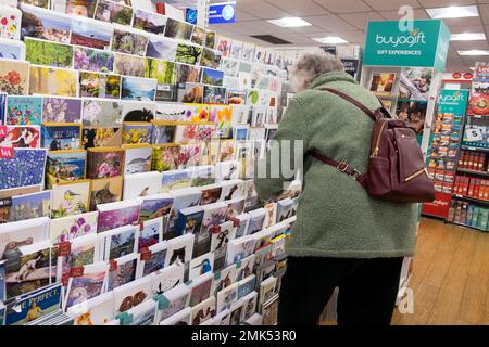 Back view older senior woman with rucksack backpack bending to look at greeting cards for sale inside stationers W.H. Smith store 28 January 2023 UK Stock Photo