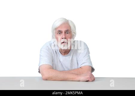 Horizontal shot of an old man sitting at a table with his arms crossed looking very surprised.  Isolated on white.  Lots of copy space. Stock Photo