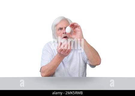 Horizontal shot of an elderly man sitting at a table examining his precious white chicken egg.  Isolated on white.  Lots of copy space. Stock Photo