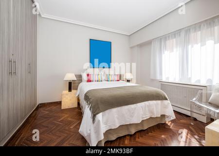 Bedroom with a double bed with colorful cushions on white pillows, a white chest of drawers with a gray blanket, a large window with white curtains an Stock Photo