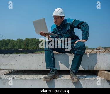 Caucasian male builder in hardhat sits on floor slabs and uses laptop at construction site.  Stock Photo