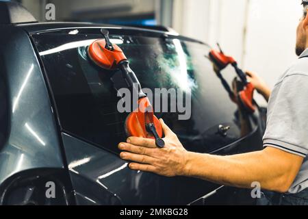 Unrecognisable man instaling rear window pane in the back of car using professional tools. Car maintenance. Garage work. Horizontal indoor shot. High quality photo Stock Photo